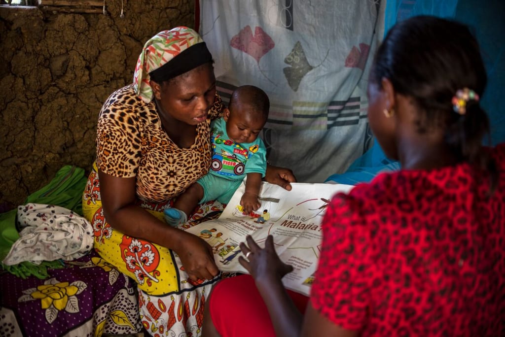 Grace is sitting in her home, wearing a brown and black animal print dress with a yellow patterned skirt. She is holding Emmanuel in her lap and talking to another woman in a red dress about malaria.